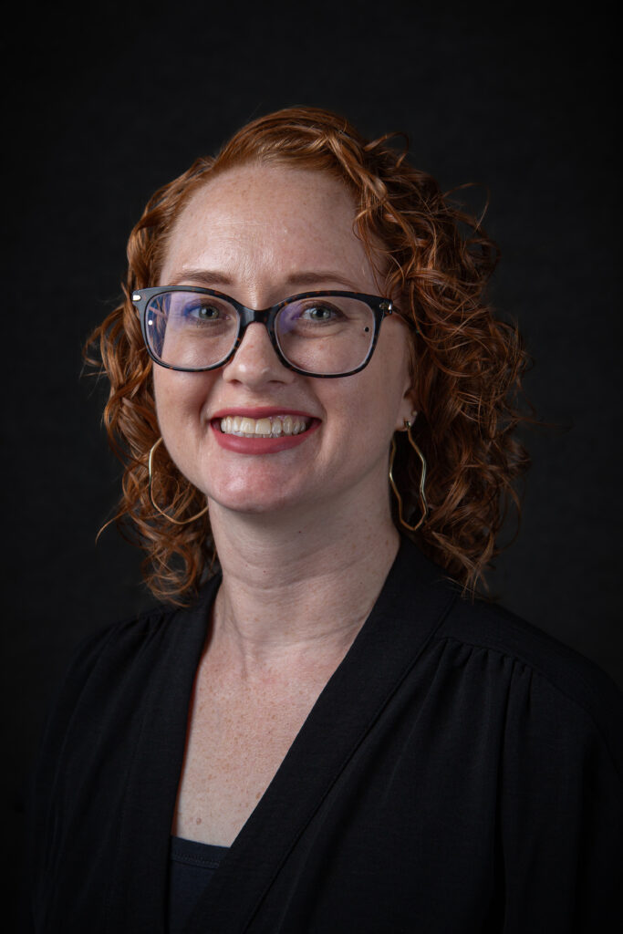 Head and shoulders photo of a smiling red headed woman wearing all black against a black background